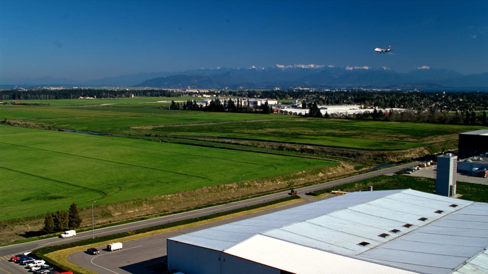 Distribution warehouses on green delta farmland with an airliner on approach overhead